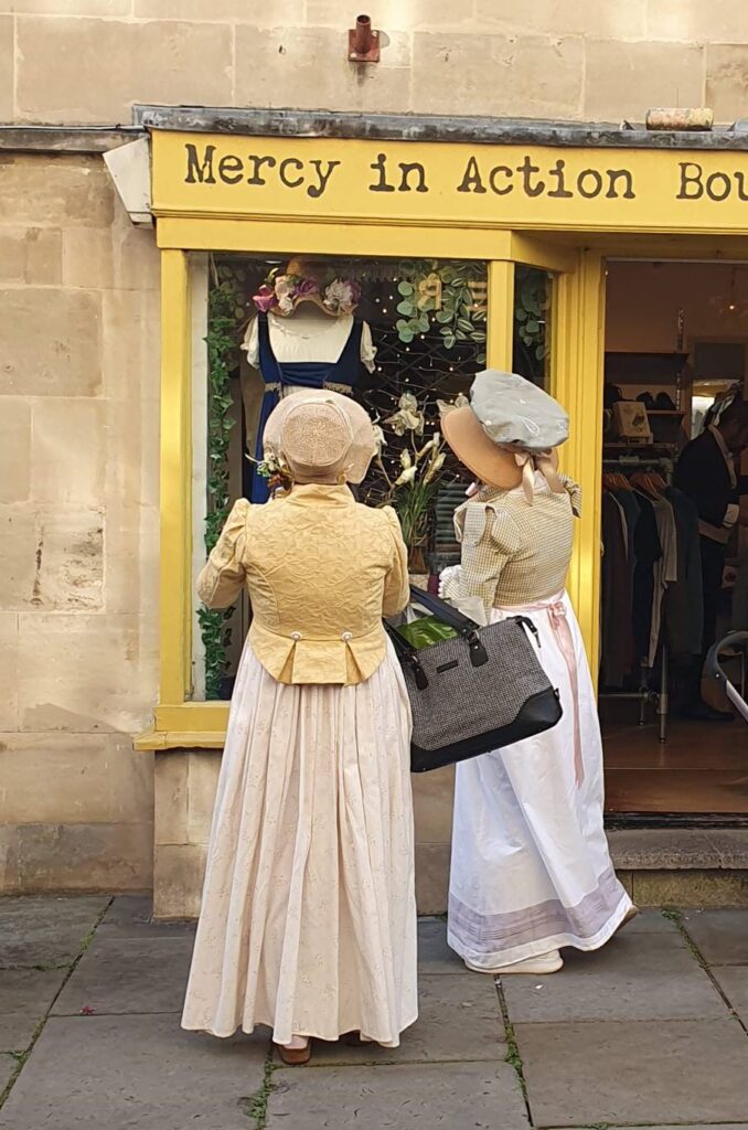 Two ladies in Regency fashions are window-shopping in Bath